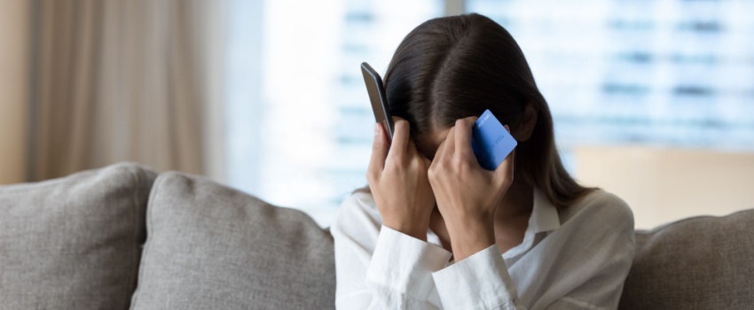 A woman holds her head with a phone in one hand and a bank card in the other.
